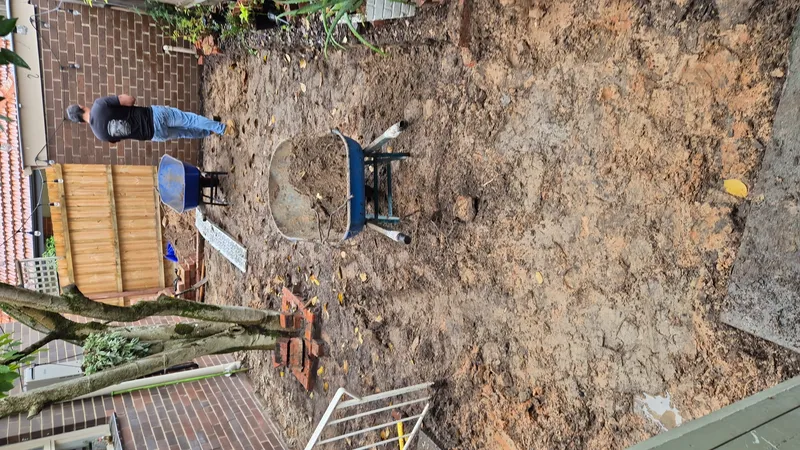 Backyard garden cleanup with blue wheelbarrow filled with soil brick wall and string lights in Melbourne